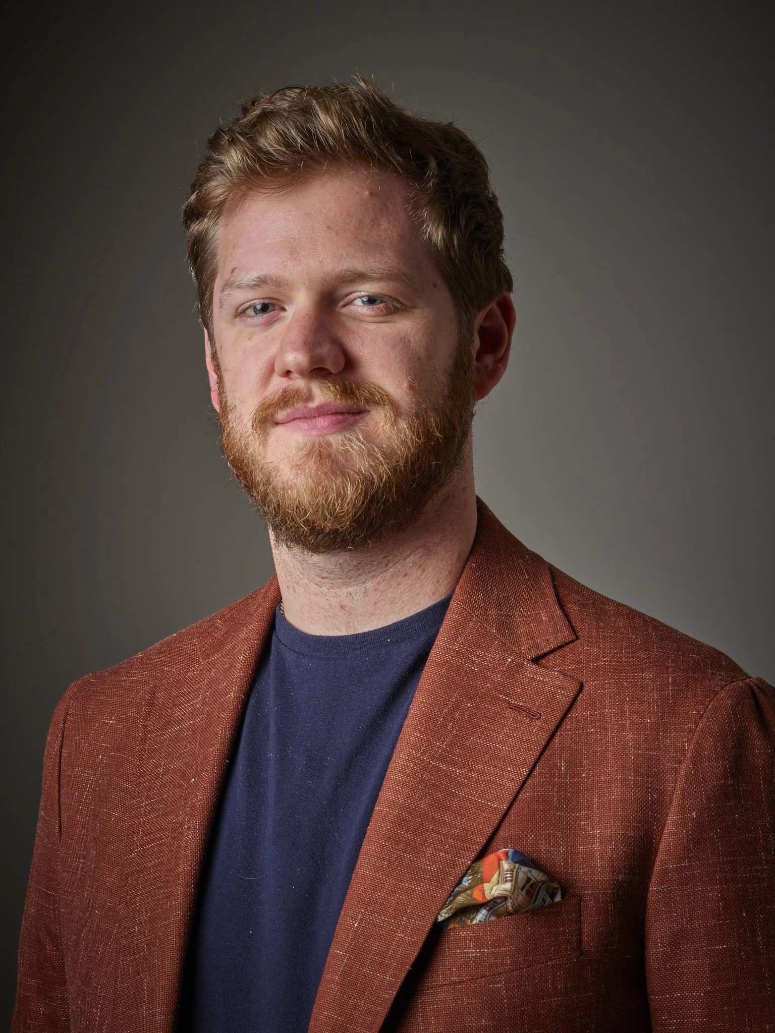Portrait of a Joshua Stephen owner and founder with blonde hair and beard wearing a rust blazer and navy shirt, against a dark gray background.