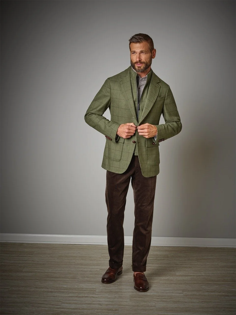 A man in a green plaid blazer, gray shirt, brown pants, and brown shoes standing against a plain background.