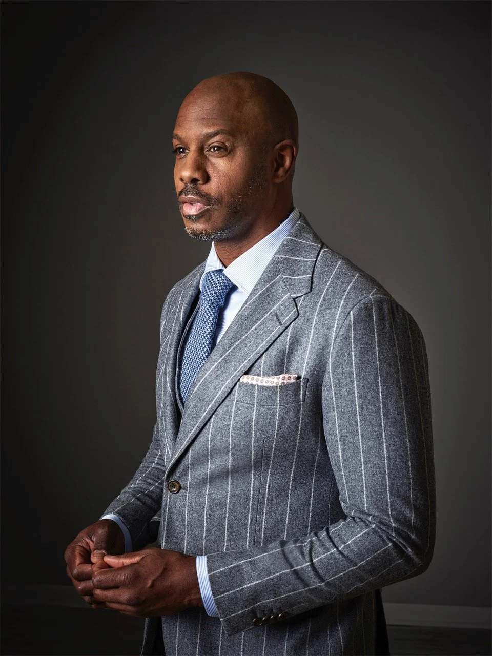 A middle-aged man wearing a grey pinstripe suit and blue tie, standing against a dark background, with a serious expression and clasped hands.
