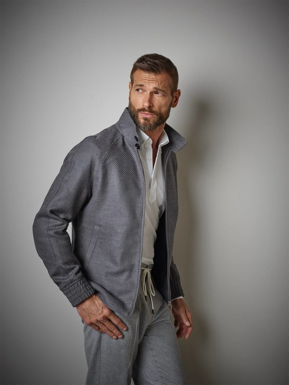 A man with a beard and short hair, wearing a gray zip-up jacket over a white shirt and gray drawstring pants, standing against a plain gray wall, looking to his left with a thoughtful expression.