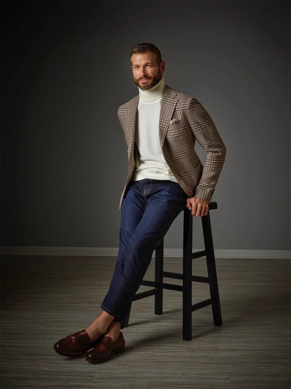 A man with a beard and styled hair sitting on a black wooden stool in a studio with a dark background, wearing a checked blazer, white turtleneck, dark jeans, and brown loafers.