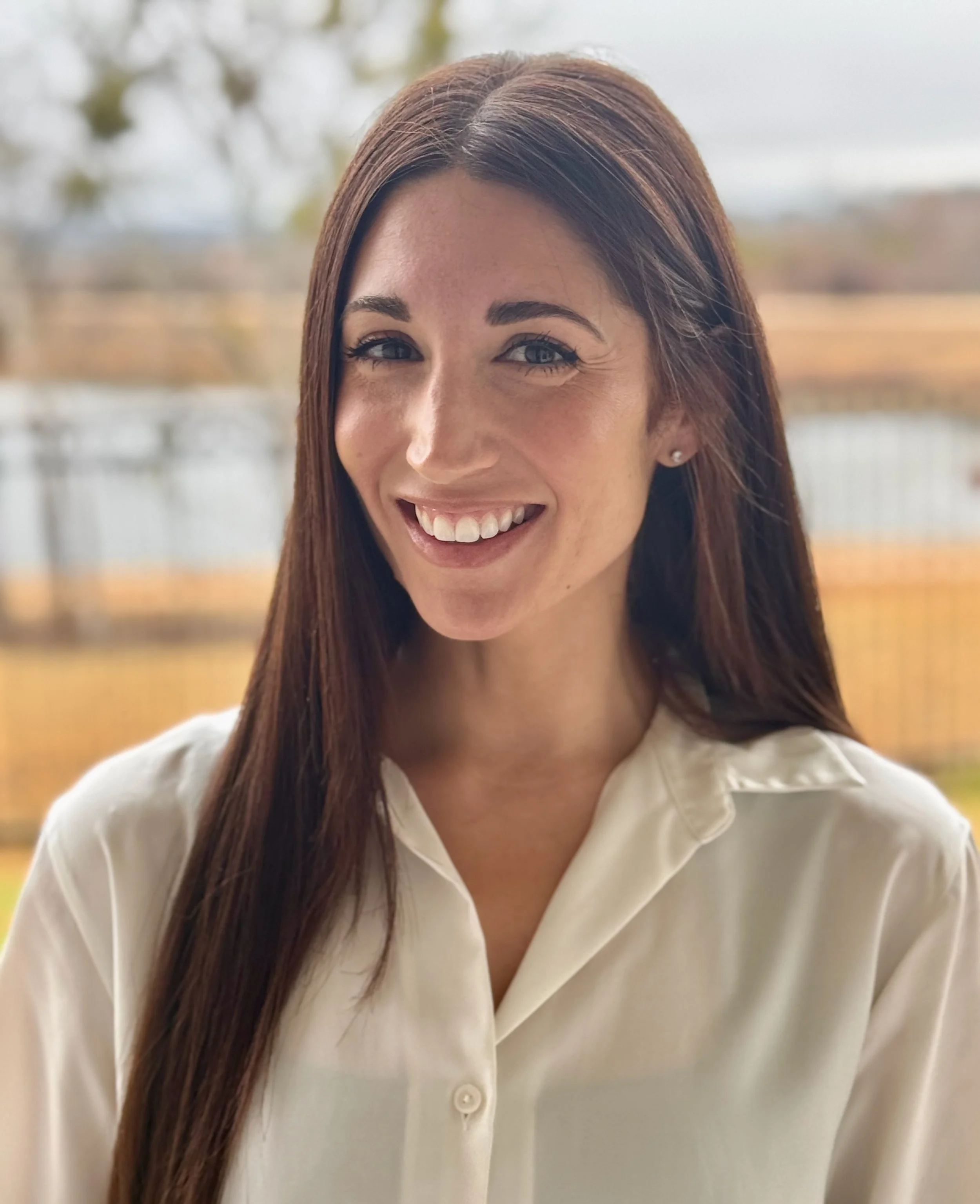 A woman with long brown hair, wearing a white blouse, smiling outdoors with blurred trees and a fence in the background.