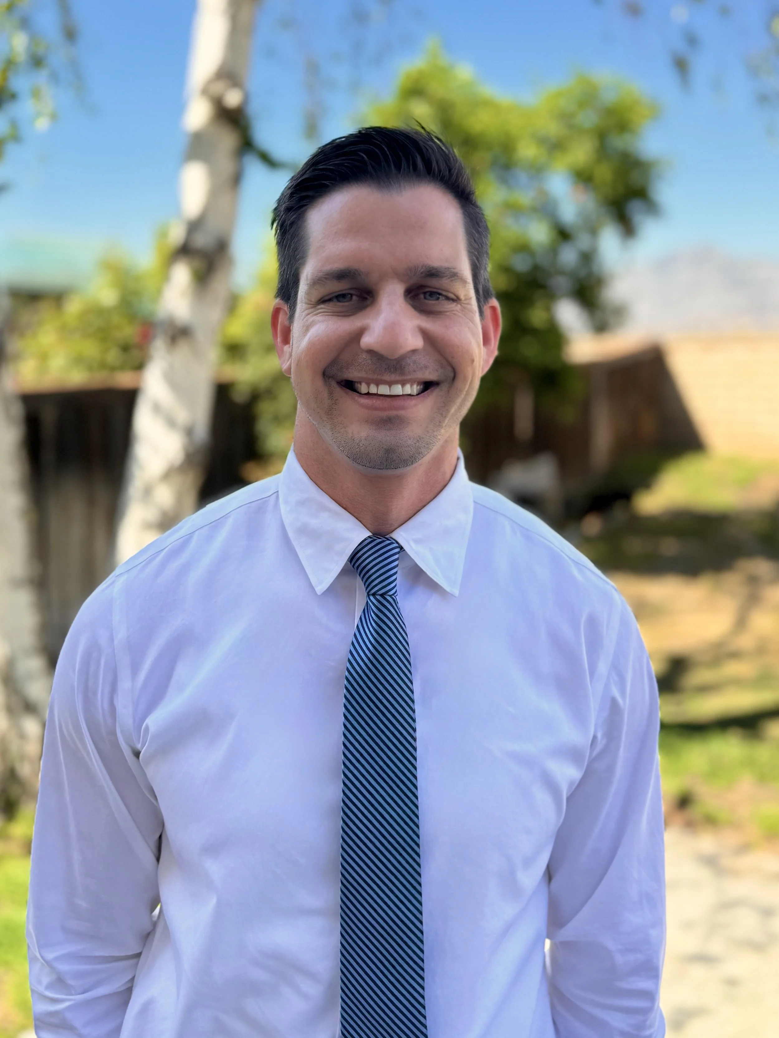 A smiling man in a white dress shirt and blue striped tie standing outdoors with trees and a wooden fence in the background.