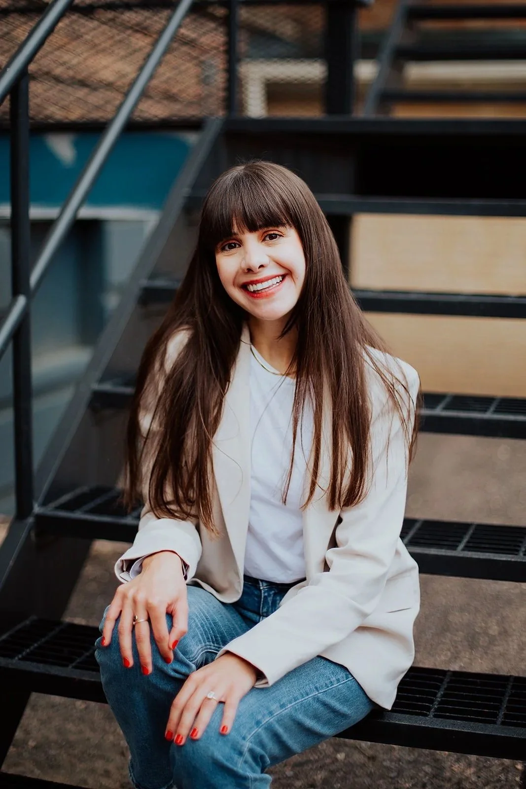 A woman with long brown hair and bangs smiling while sitting on outdoor metal stairs, wearing a white blazer, white t-shirt, and blue jeans.