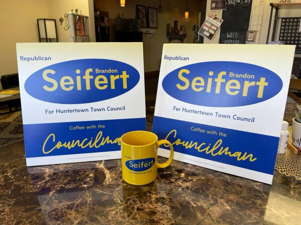 Political campaign signs and a coffee mug promoting Brandon Seifert for Huntertown Town Council, displayed on a marble table.