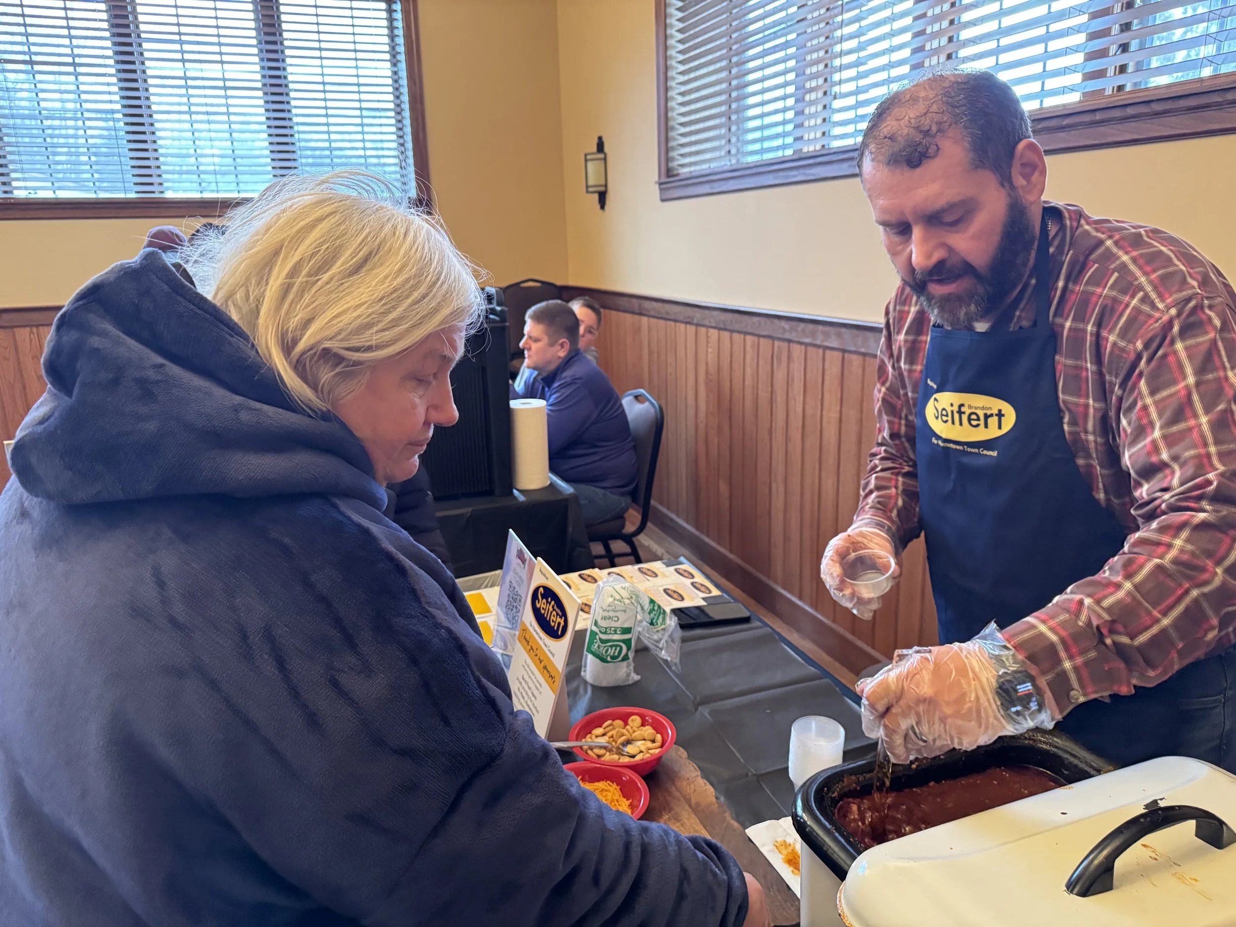 A man Brandon Seifert serving food from a crockpot to a woman at a table in a restaurant or community center. The man is wearing a blue apron Chili competition Cookoff. Huntertown, Indiana.