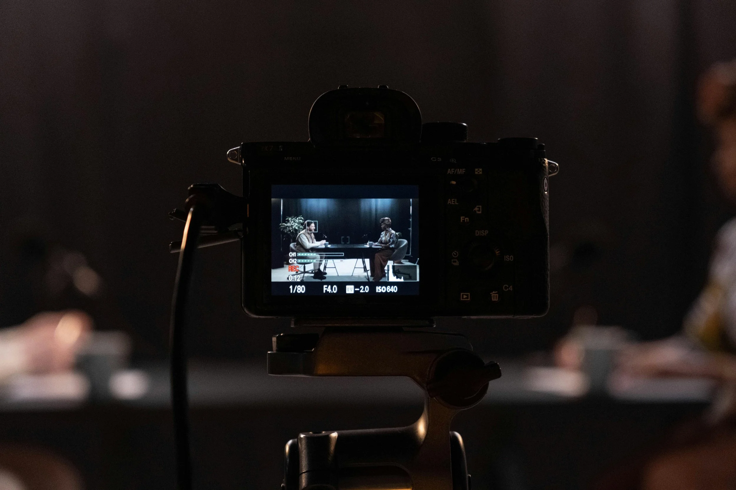 A camera recording two women during an interview set in a studio with dark background, with one woman wearing a white blazer and the other in patterned clothing, seated across from each other at a table.
