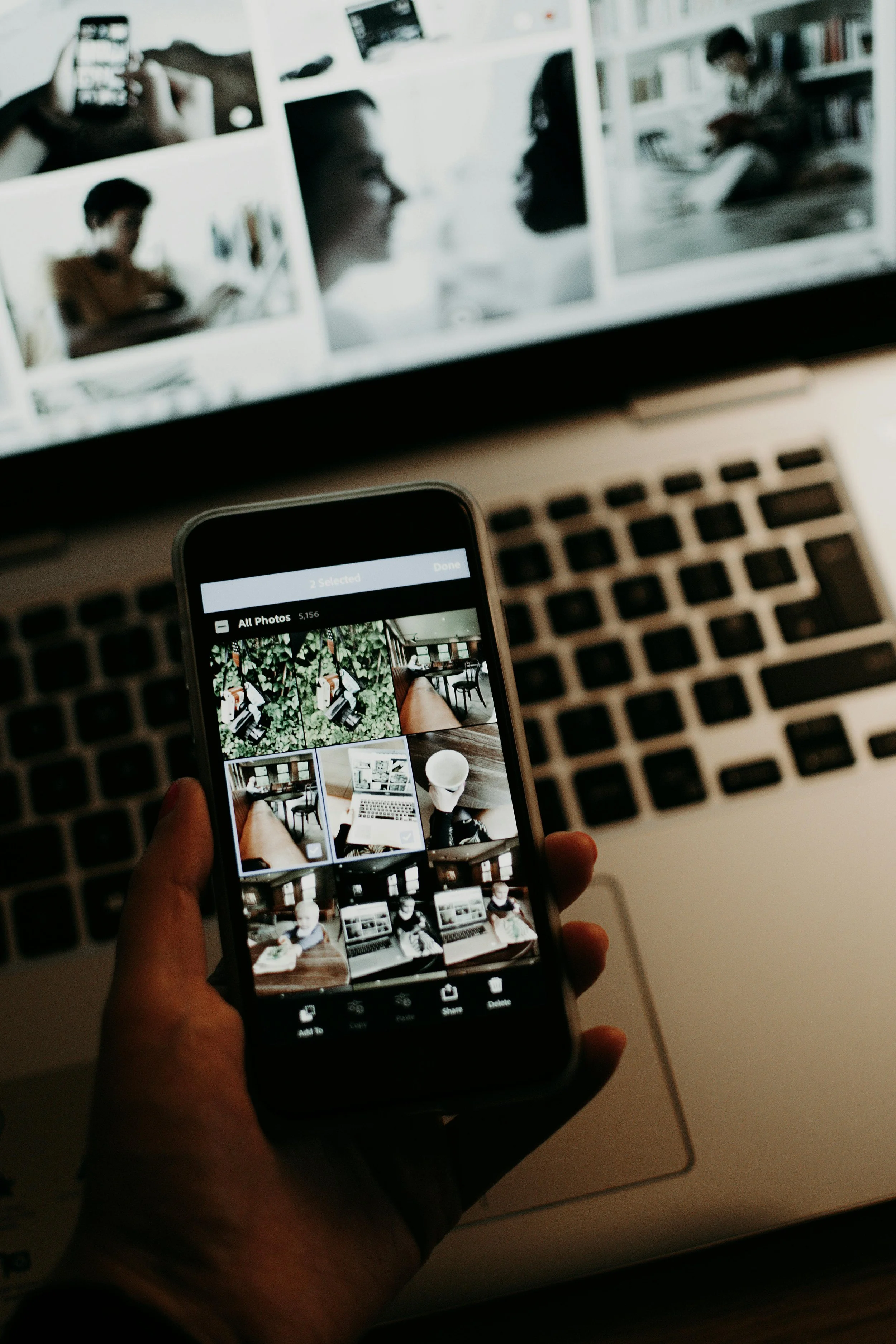 Close-up of a hand holding a smartphone displaying photo thumbnails, with a laptop in the background showing a collage of black-and-white portrait photos, some of people reading and working.
