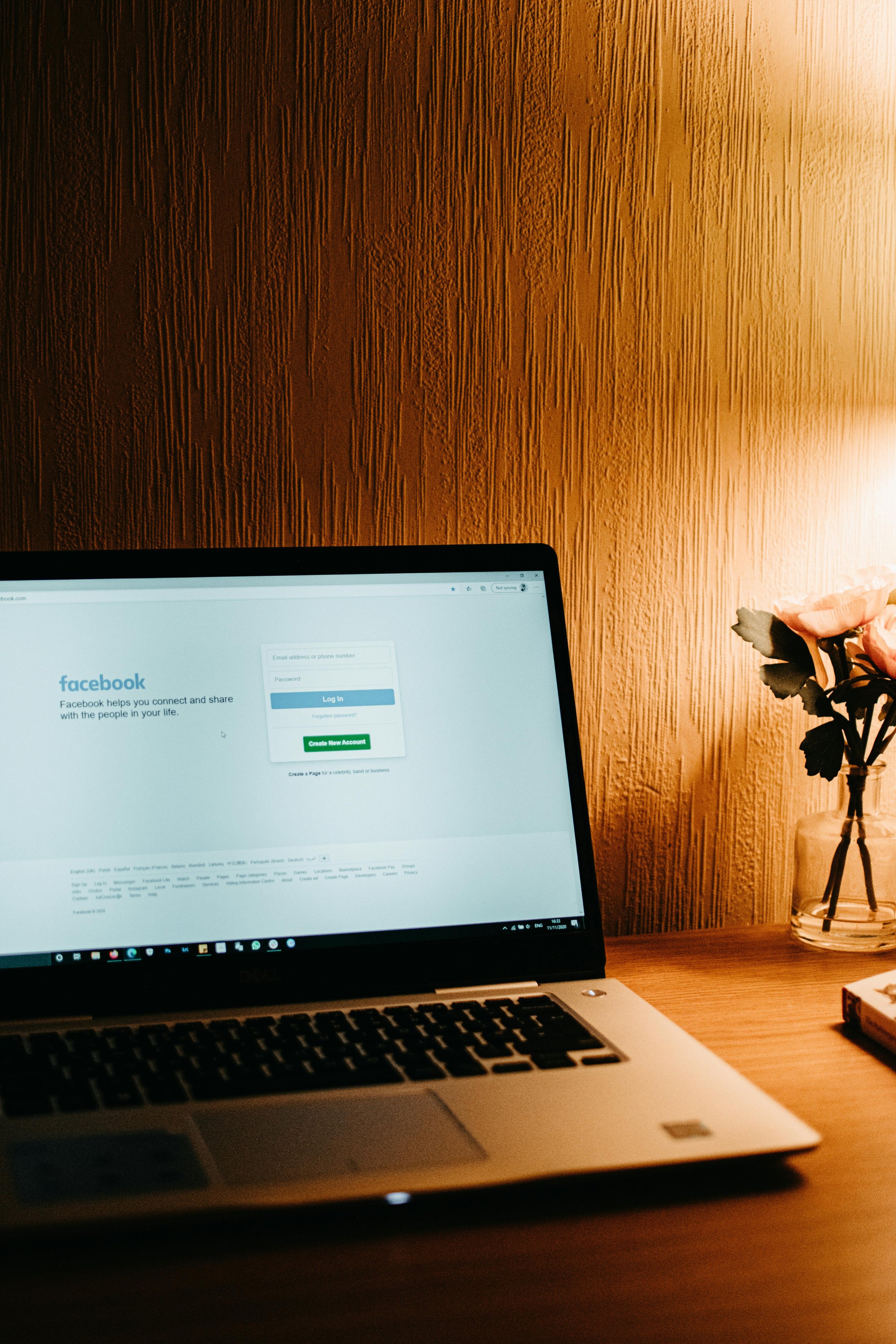 A laptop on a wooden desk displaying the Facebook login page, with a vase of pink roses to the right, against a textured wooden wall with warm lighting.