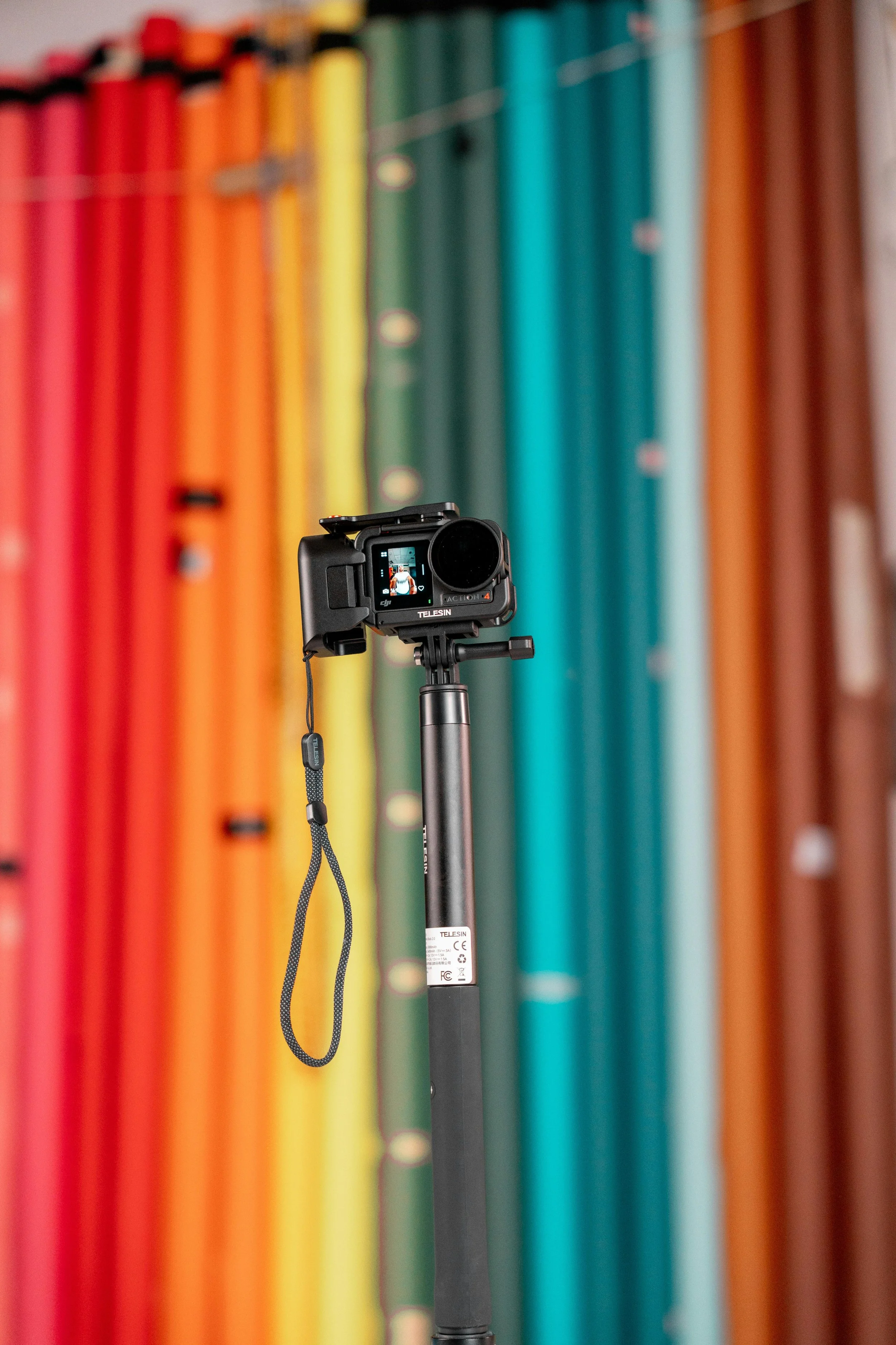 A digital camera mounted on a black tripod, pointed at a colorful background of vertical fabric rolls in rainbow colors.