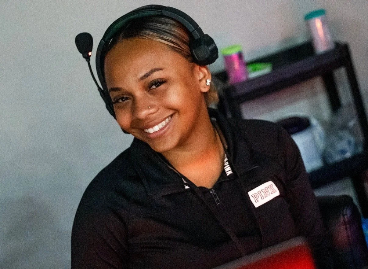 A smiling woman with a headset, earrings, and a black jacket, sitting indoors with a shelf in the background.