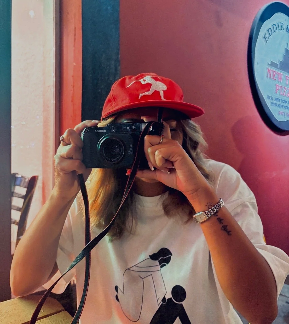 Person taking a selfie with a Canon camera, wearing a red baseball cap with a white lion logo, white t-shirt with a black outline of a person with sunglasses, jewelry including bracelets and a ring, sitting inside a restaurant with pink walls and a sign for a pizza place in the background.