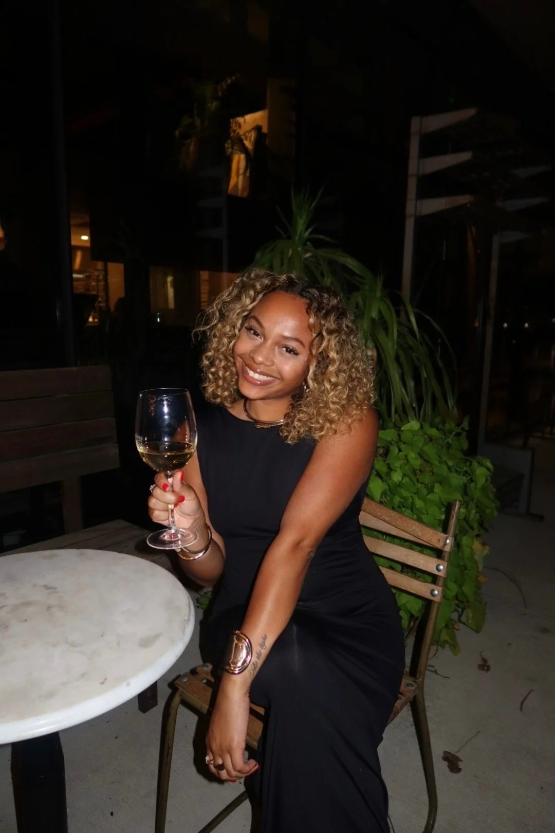 A woman with curly blonde hair, wearing a black sleeveless dress, sitting outdoors at night, holding a glass of white wine, smiling at the camera.