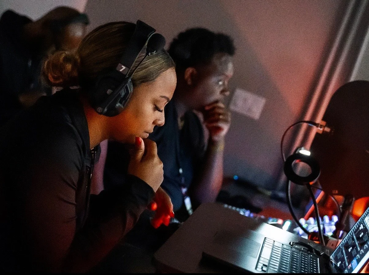 Two people working at a computer station in a dark environment, wearing headphones and focusing on their screens.