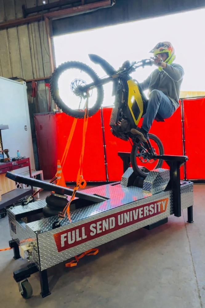 Person wearing a helmet and gray hoodie riding a yellow and black electric dirt bike on a roller coaster-like track in a garage or workshop. The lift platform has 'FULL SEND UNIVERSITY' written on it.
