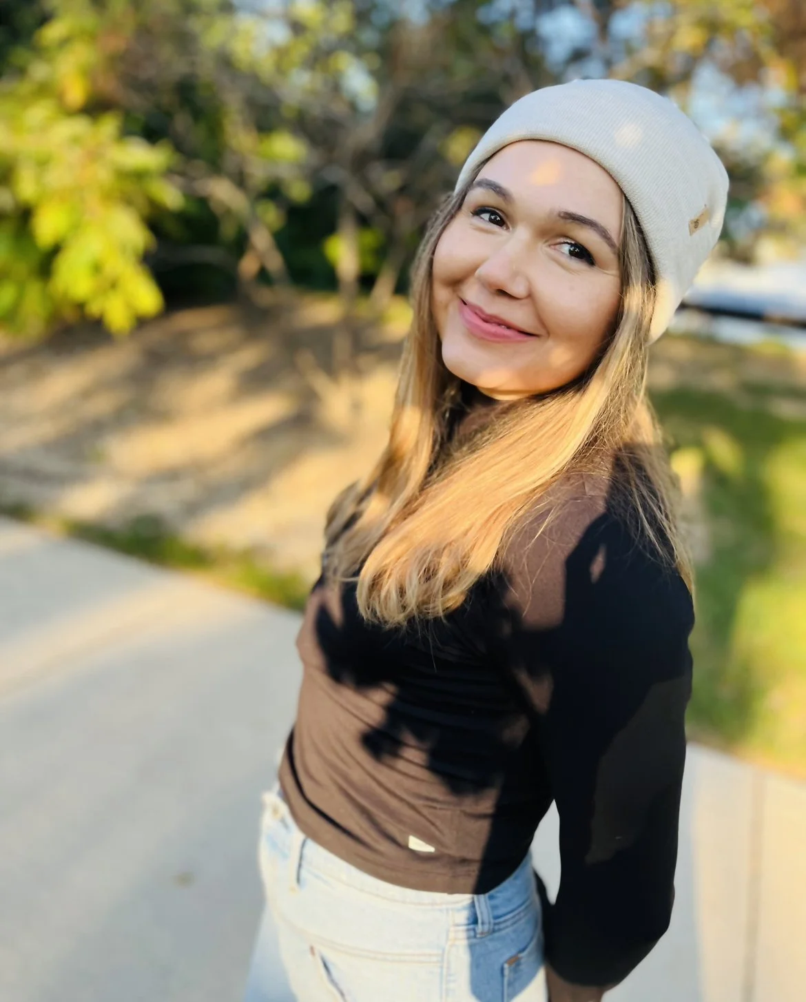 Smiling young woman with long blonde hair, wearing a beige beanie and a black jacket, standing outdoors with blurred trees and sunlight in the background.