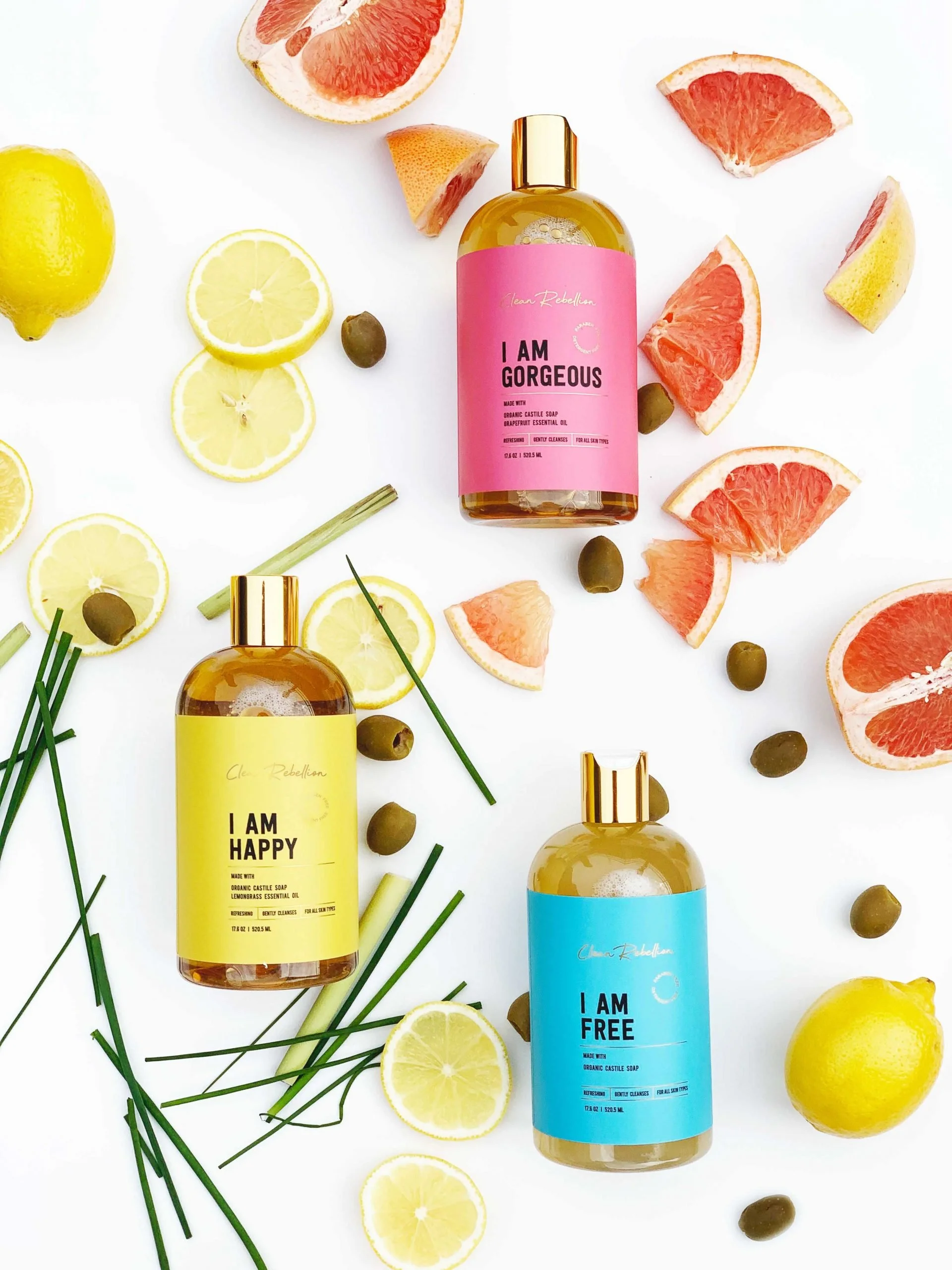 Three bottles of soap labeled 'I AM GORGEOUS,' 'I AM HAPPY,' and 'I AM FREE,' surrounded by lemon, grapefruit, and olive slices, along with green herbs, on a white background.