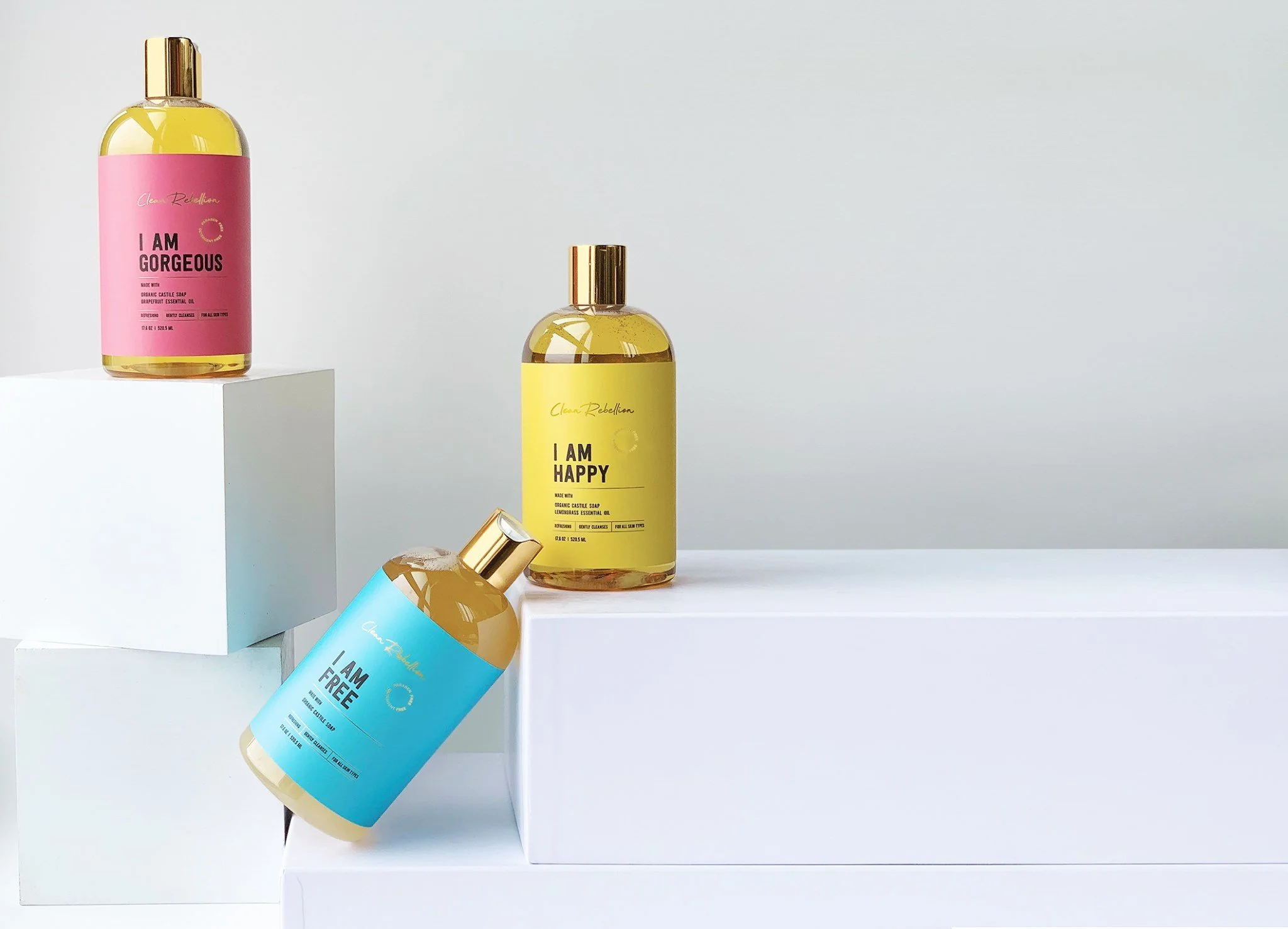 Three colorful bottles of body wash with labels reading 'I AM GORGEOUS,' 'I AM HAPPY,' and 'I AM FREE,' arranged on white geometric platforms against a plain background.