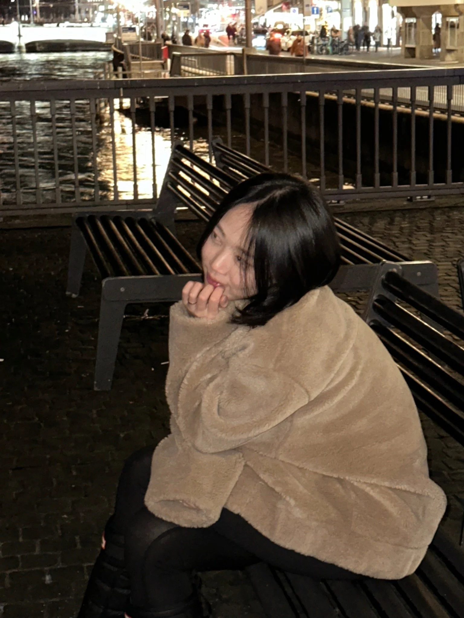A woman with black hair sitting on a bench near a waterfront at night, wearing a beige fuzzy coat and smiling while resting her face on her hand.