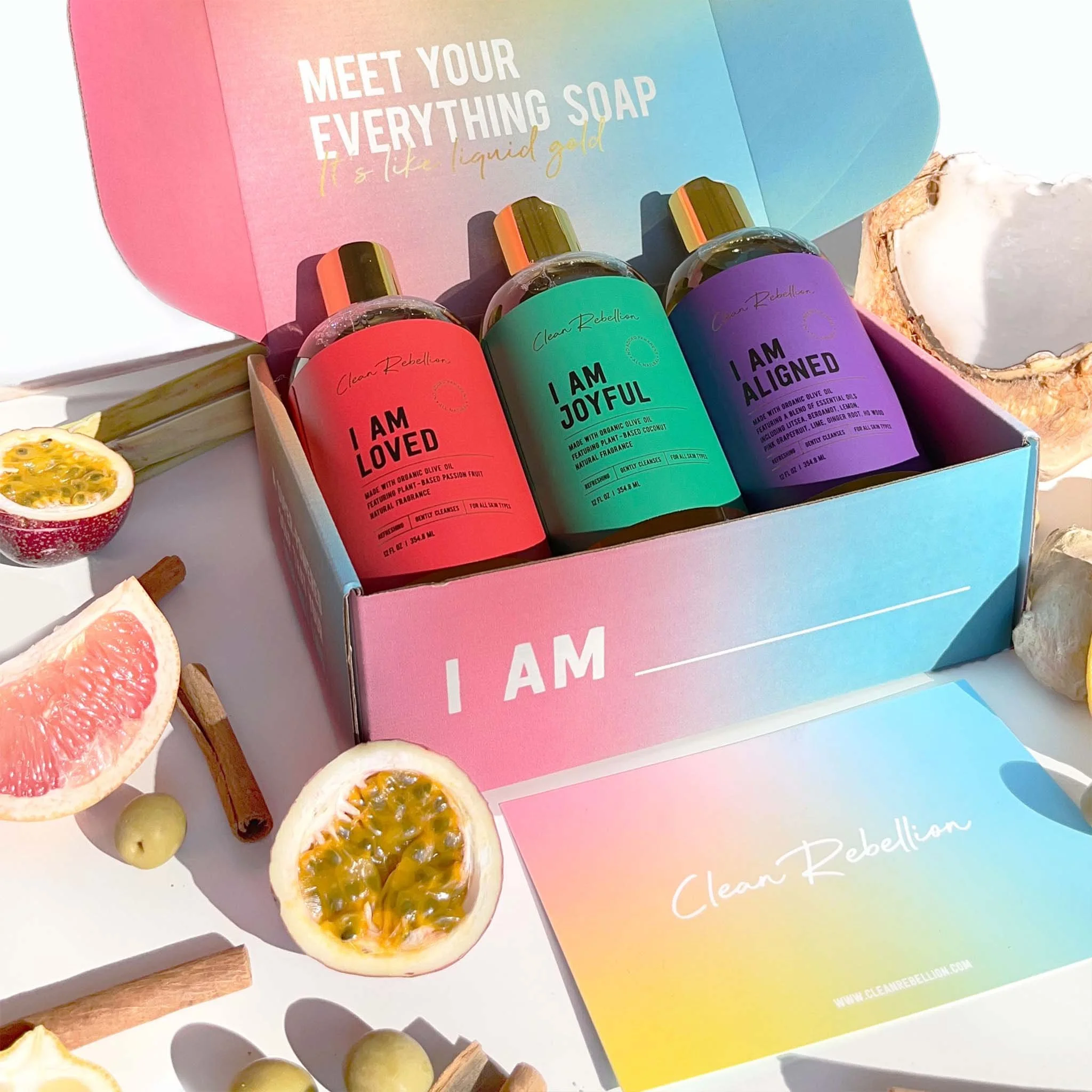 Colorful box containing three bottles of liquid soap labeled 'I AM LOVED,' 'I AM JOYFUL,' and 'I AM ALIGNED.' The box has a pastel gradient design with the phrase 'MEET YOUR EVERYTHING SOAP' on the interior lid. Surrounding the box are slices of passion fruit, grapefruit, cinnamon sticks, and green grapes, set against a white background with bright lighting.