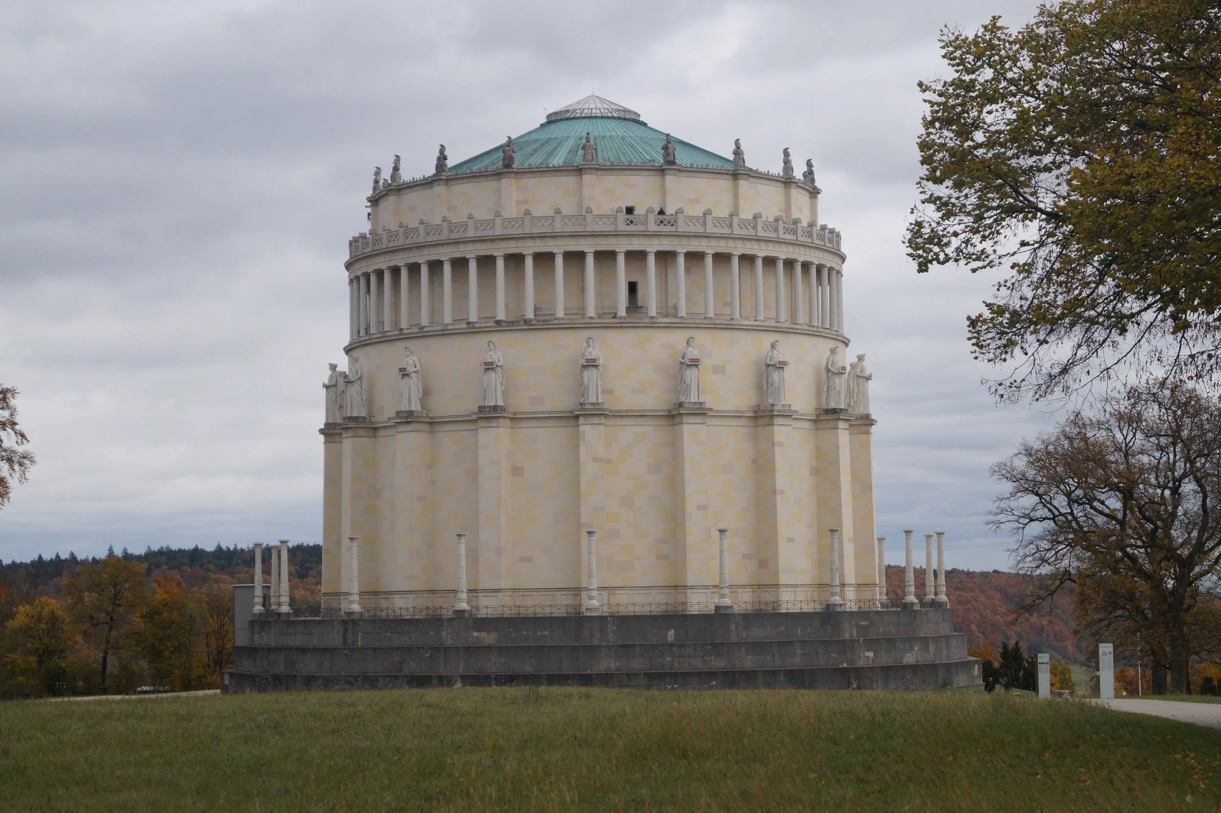Der Himmel ist bewölkt, und im Vordergrund befindet sich ein weißer Rundbau mit mehreren Säulen und Statuen auf der Dachlinie, umgeben von Bäumen mit Herbstlaub.