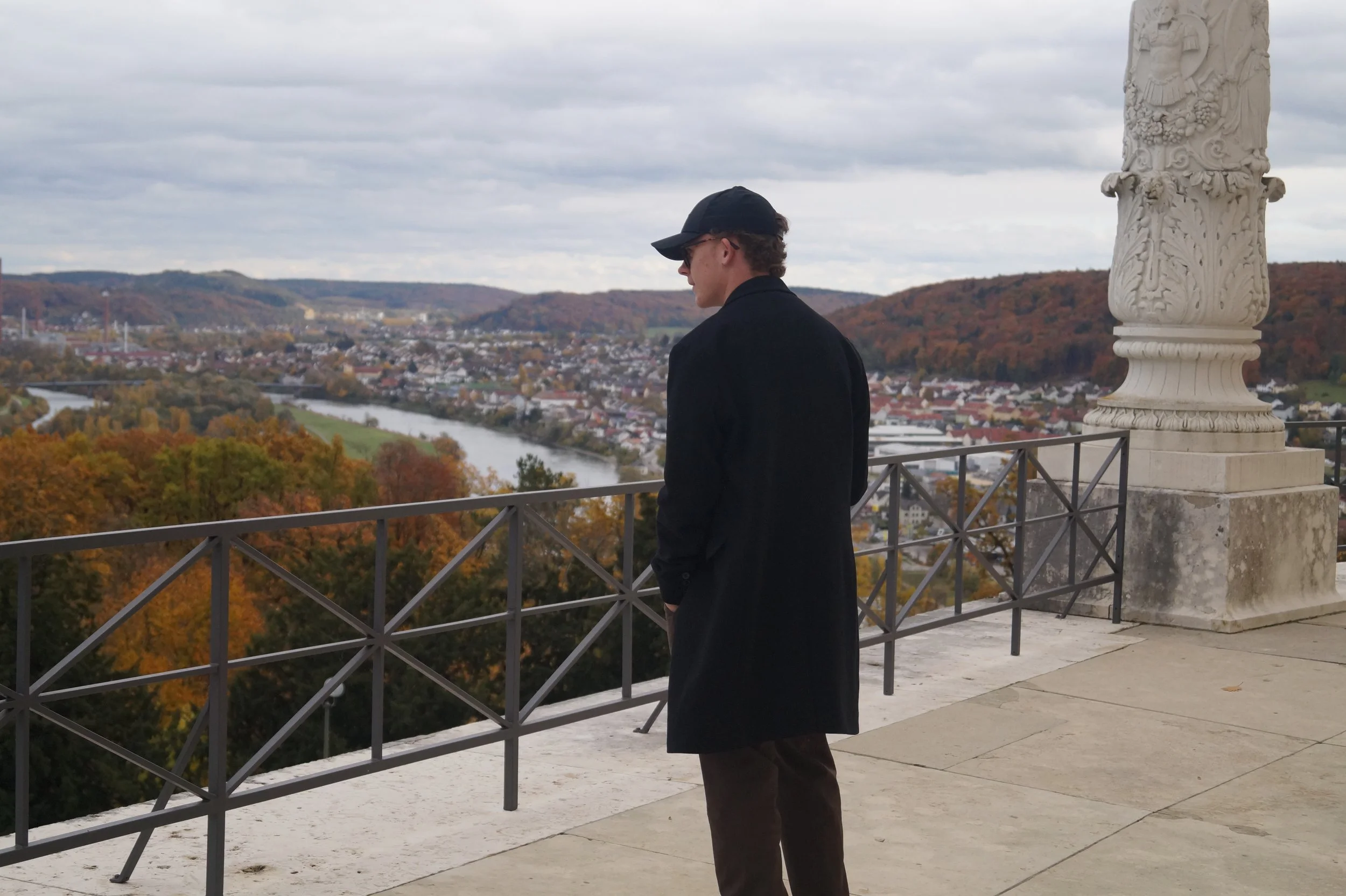 Ein junger Mann steht am Aussichtspunkt mit Blick auf eine Stadt, Fluss und Hügel im Hintergrund, trägt schwarze Jacke, Mütze und Sonnenbrille, bei bewölktem Himmel.
