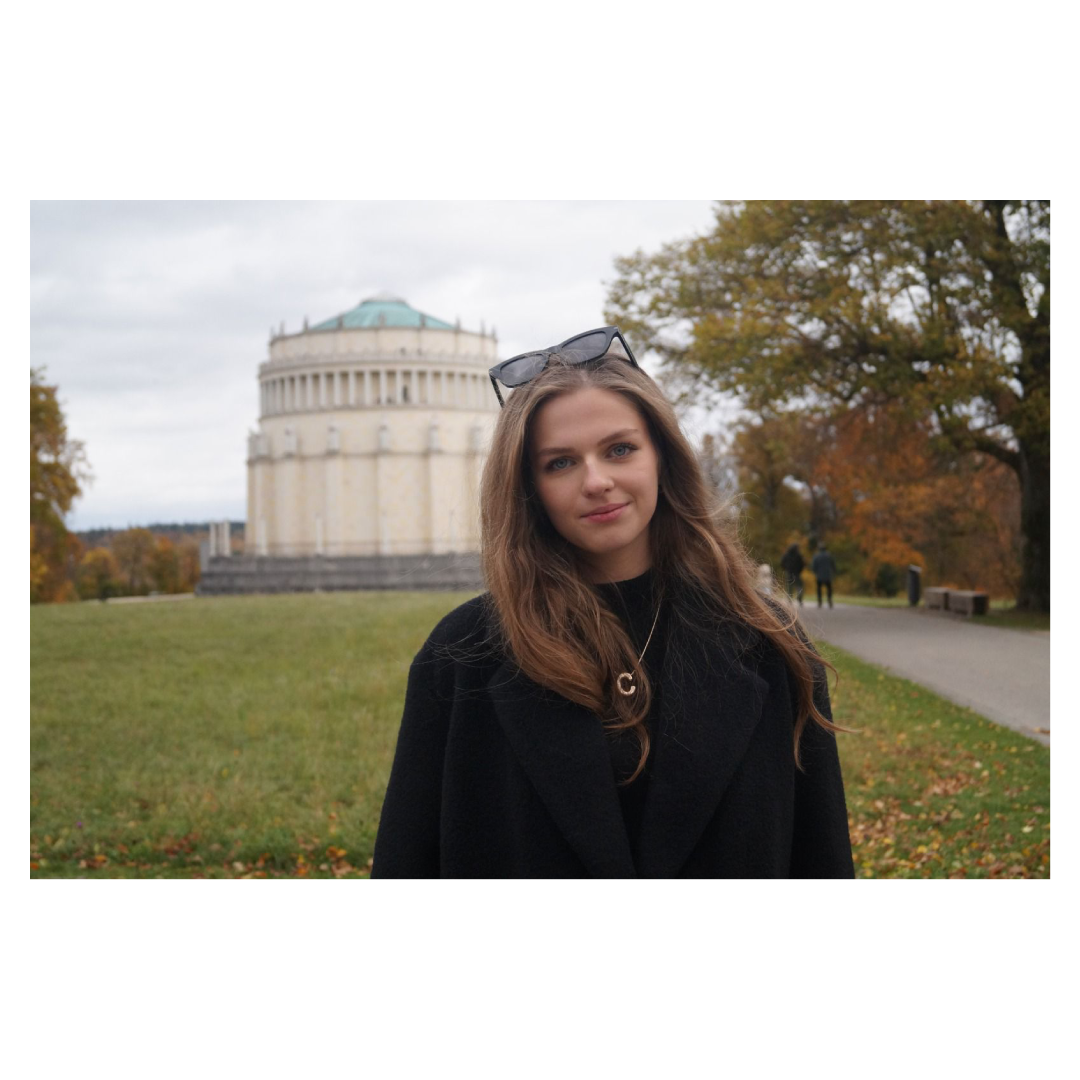 Ein junges Mädchen steht vor einer historischen Kirche im Park, mit Herbstbäumen im Hintergrund.