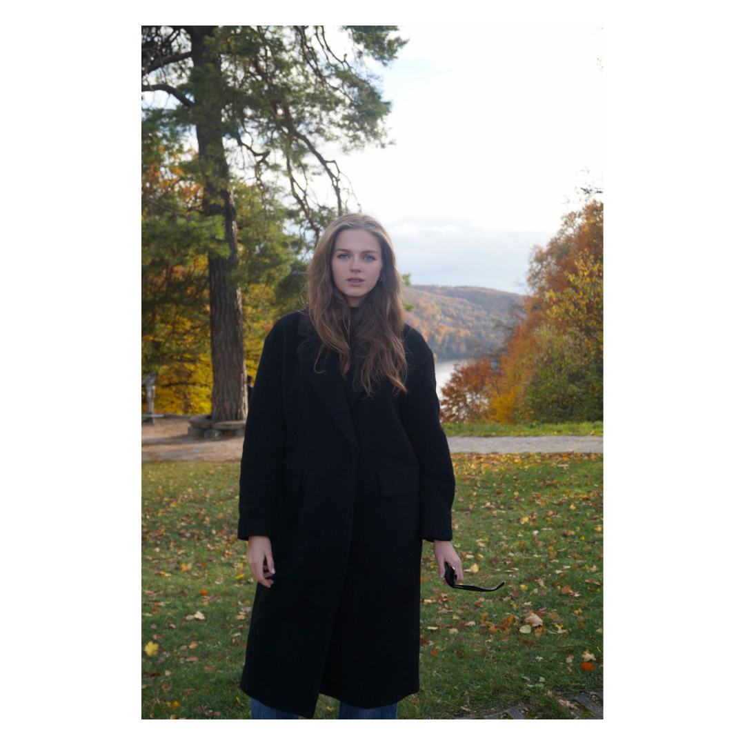 Junge Frau mit langen braunen Haaren im schwarzen Mantel steht im Park im Herbst, mit bunten Blättern auf dem Boden und Bäumen im Hintergrund.