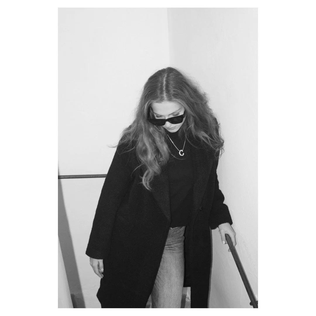 Eine junge Frau mit langen, lockigen Haaren, dunkler Sonnenbrille, schwarzer Jacke, grauen Jeans, auf einer Treppe, schwarz-weiße Aufnahme.