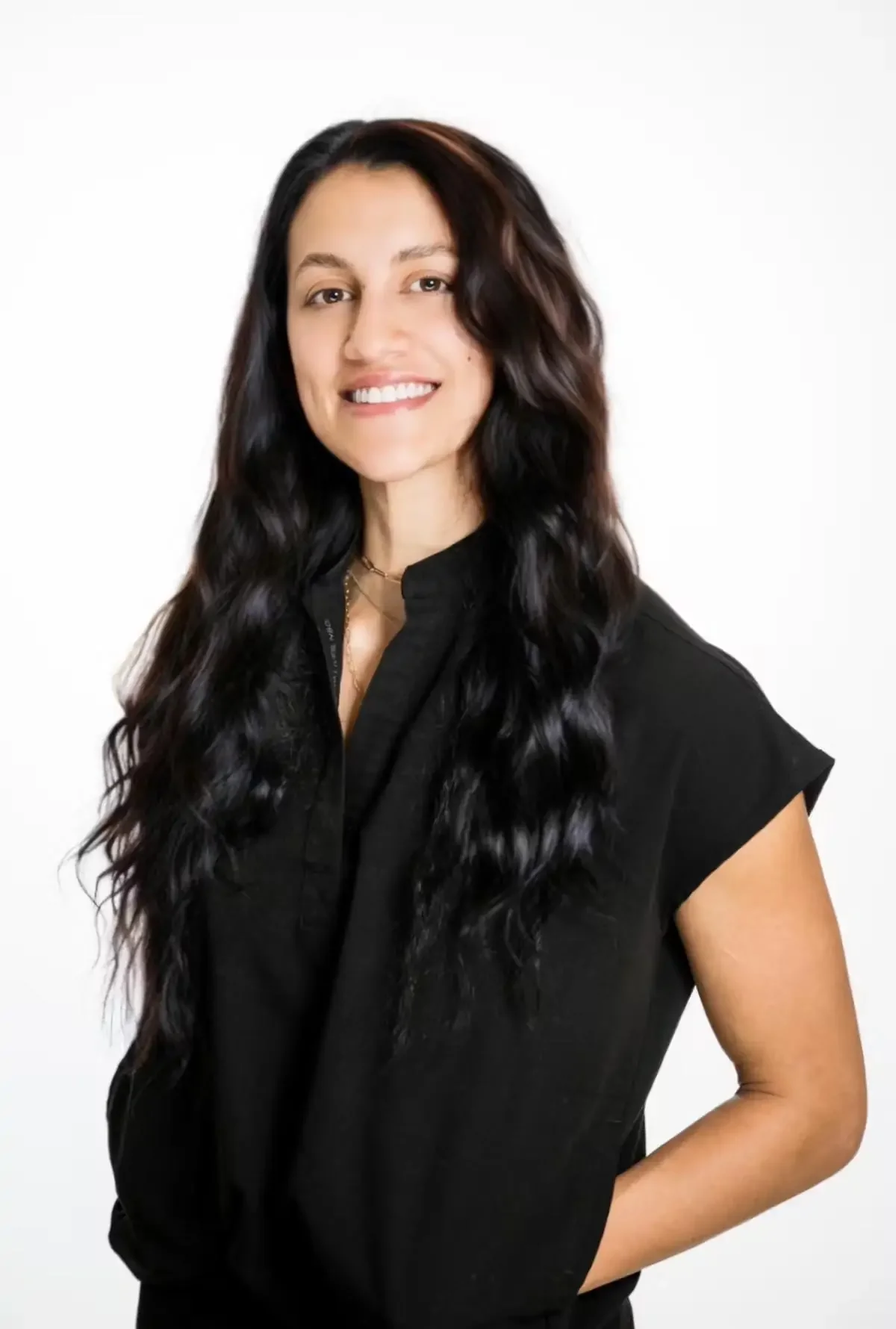 A woman with long, wavy black hair, smiling, wearing a black short-sleeved blouse, standing against a white background.