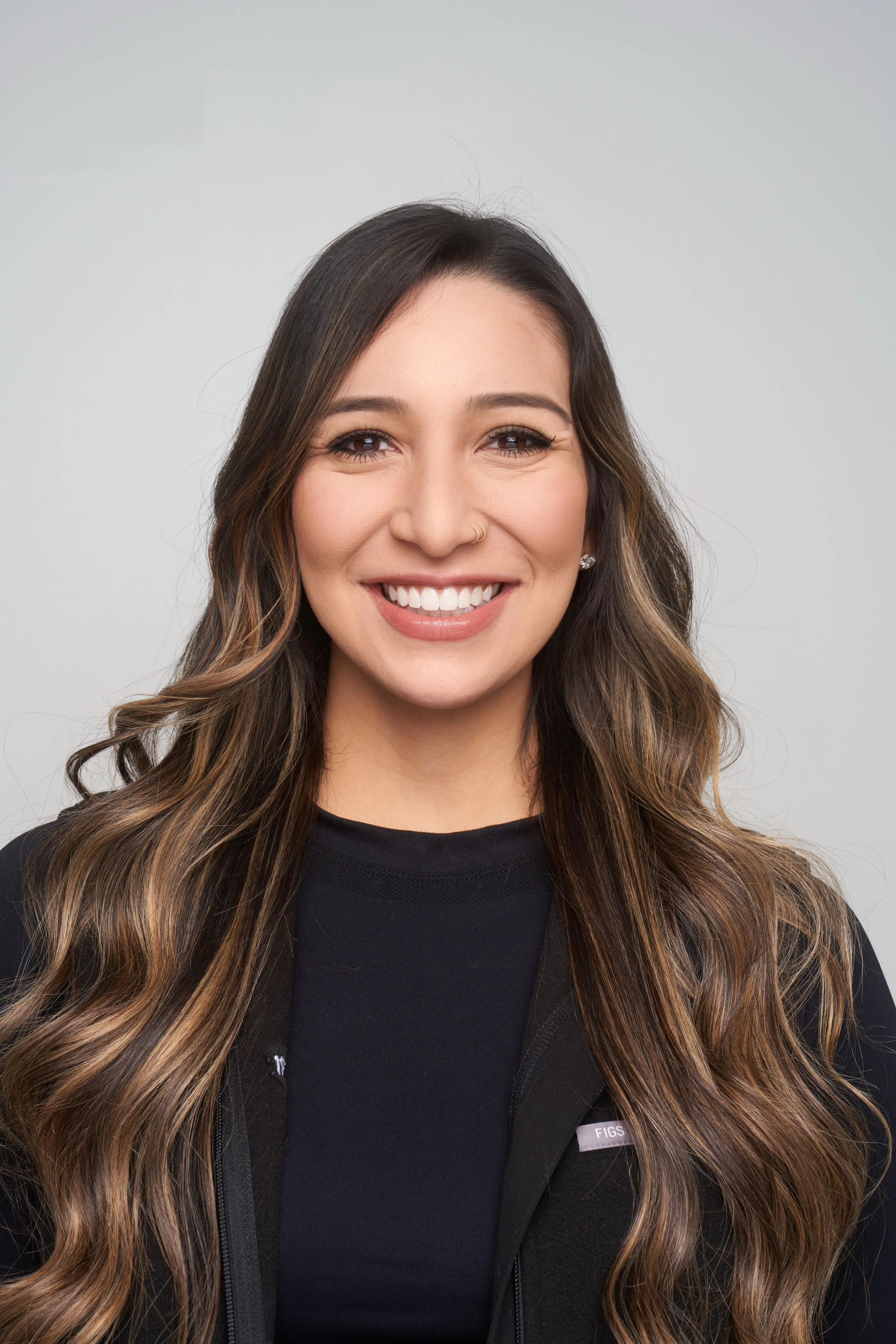 A woman with long wavy brown hair, smiling, wearing a black top and a black jacket, against a plain light gray background.