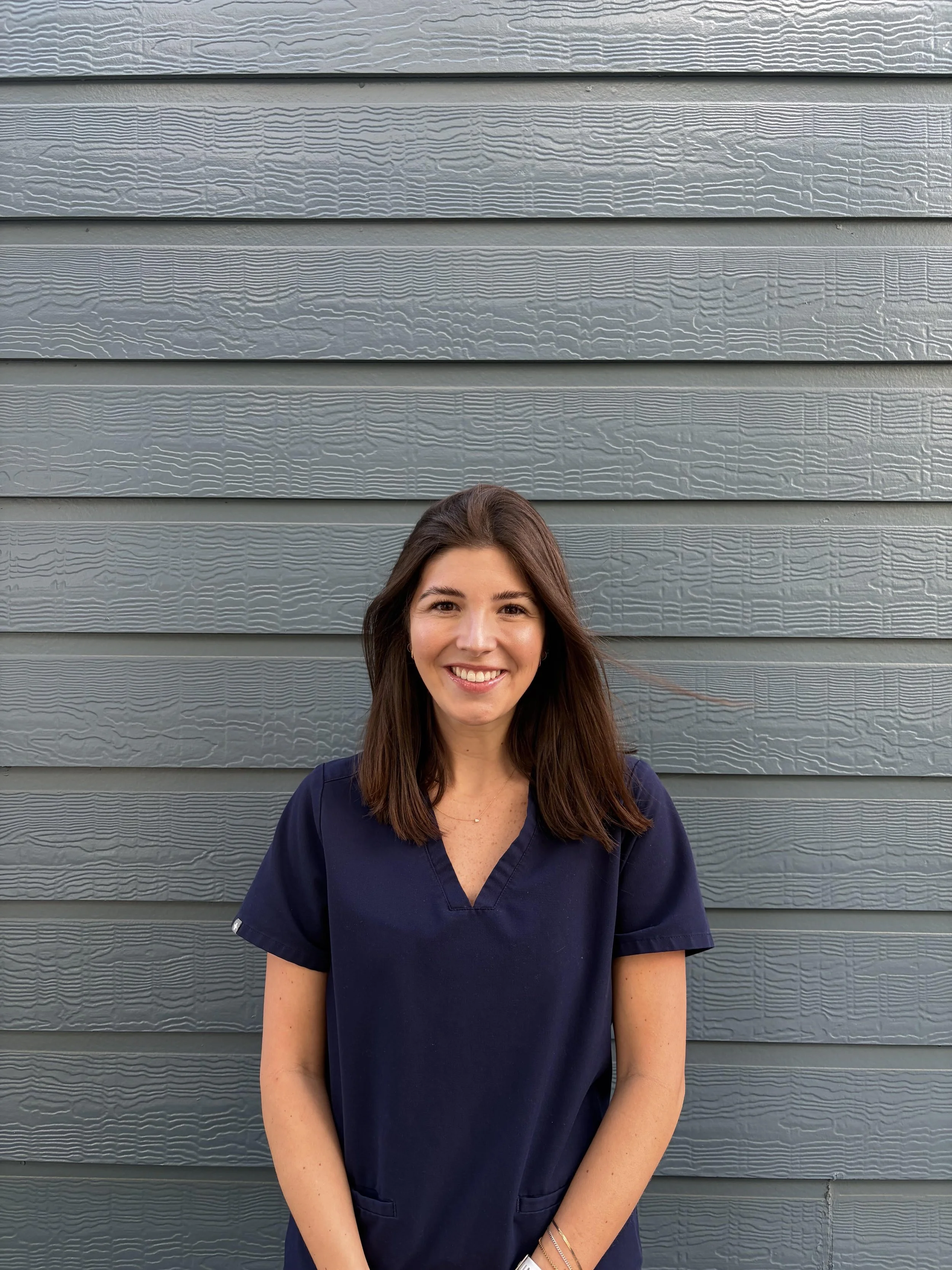 A woman with shoulder-length brown hair, wearing a navy blue scrub top, smiling and standing in front of a textured gray wooden wall.