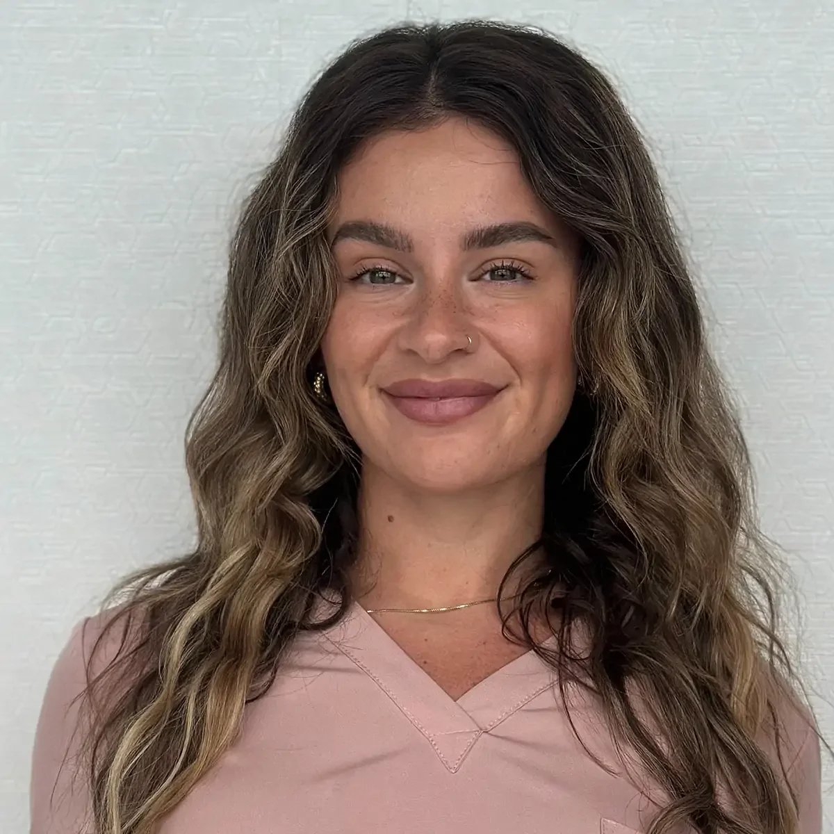 A woman with long, wavy brown hair, a nose ring, and light eye makeup, smiling in front of a plain textured wall, wearing a light pink V-neck top and gold jewelry.