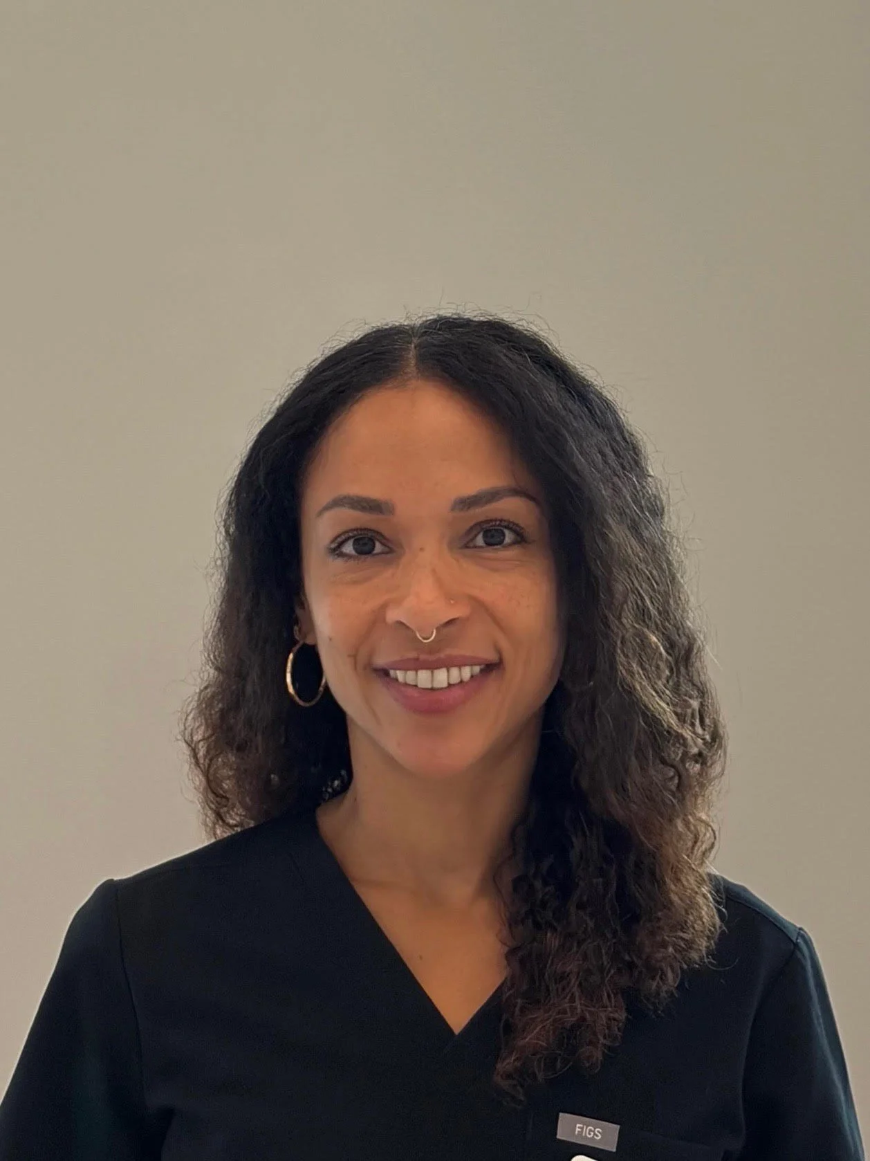 A woman with dark curly hair, wearing hoop earrings, a septum piercing, and a black top, smiling at the camera against a plain light-colored background.
