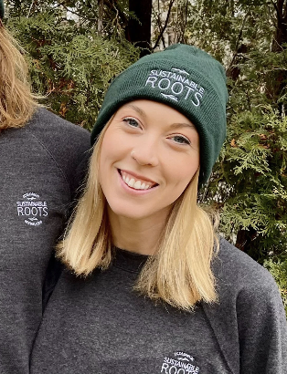 Young woman smiling outdoors wearing a green beanie and a dark gray jacket, both with a sustainable roots logo.