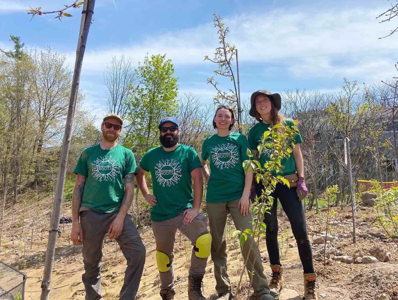 Four people outdoors, standing among small trees and plants, wearing matching green shirts with a tree logo and words 'Sustainable Roots,' smiling, under a blue sky with some clouds.