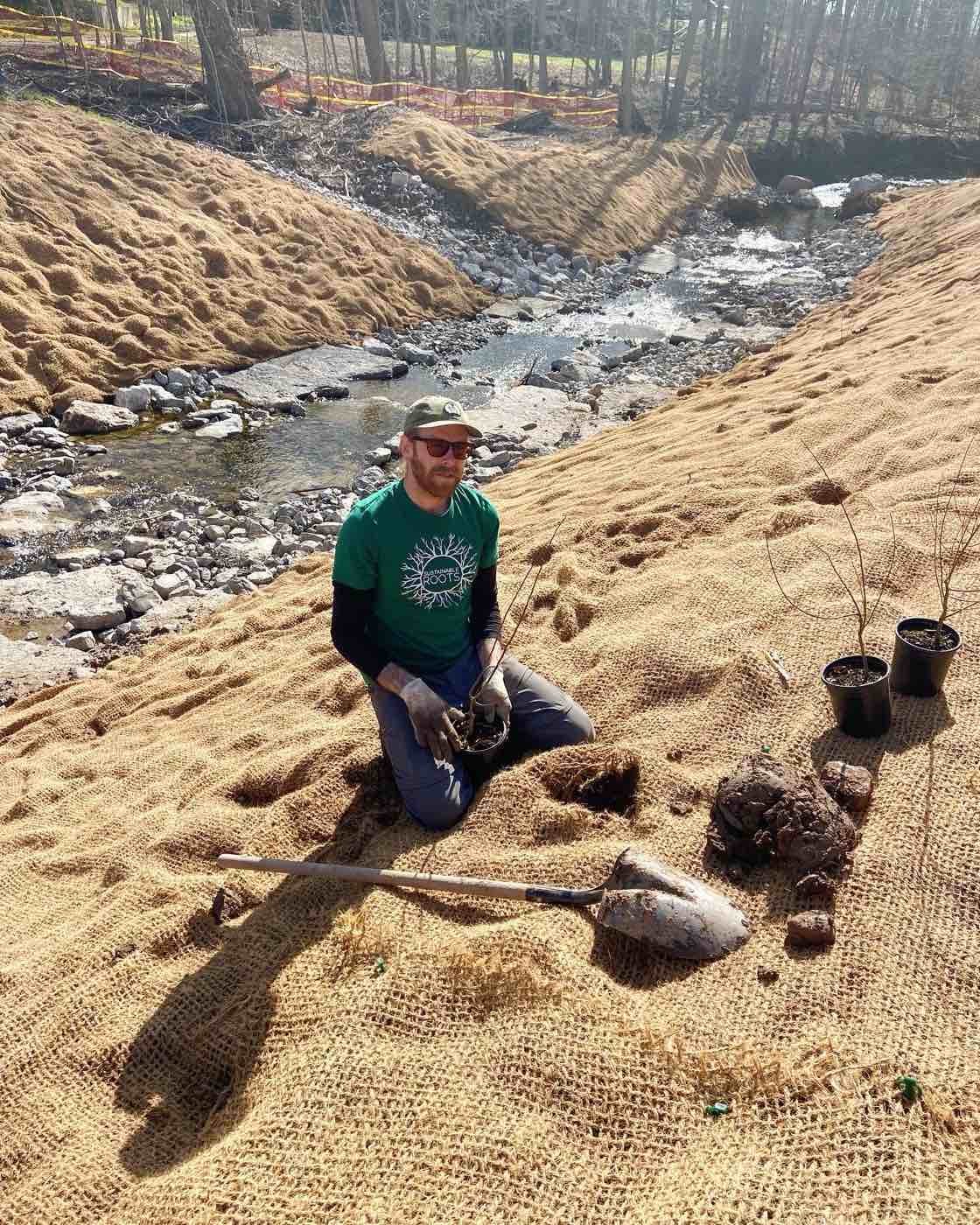 A man kneeling on a burlap-covered slope by a stream, holding a metal detector near a large rock, with planting pots nearby. Trees and a fenced area are in the background.