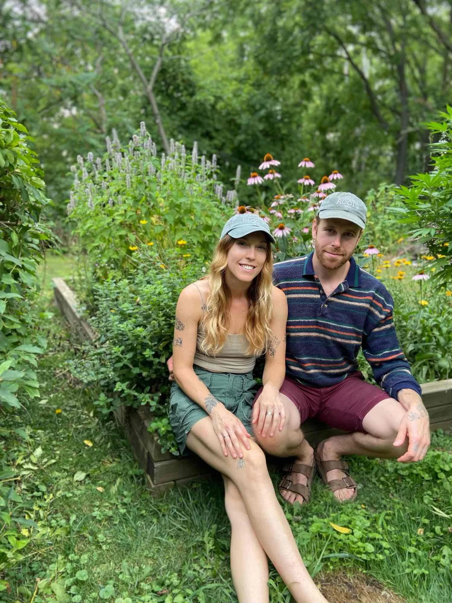 A young woman and a young man sitting on a wooden bench in a lush green garden, both wearing grey caps, with flowering plants and trees in the background.