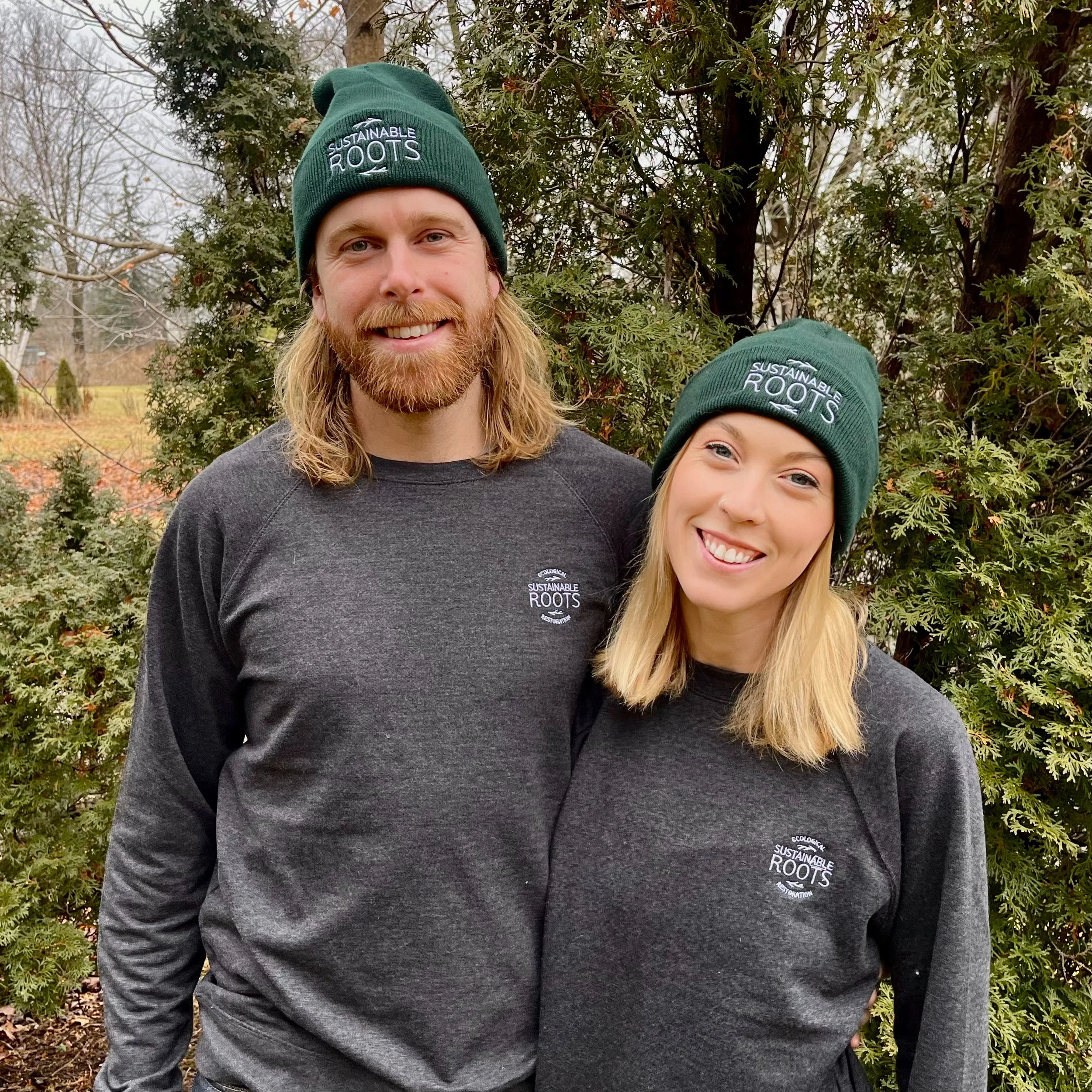 Close-up of two smiling people wearing green beanies with the logo 'Sustainable Roots'. One person has long blonde hair and a beard, and the other has long blonde hair and a nose piercing. They are standing outdoors in front of a bush.