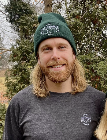 A man with long blonde hair and a beard wearing a green beanie that says 'Sustainable Roots' and a dark gray long-sleeve shirt, standing outdoors among trees.