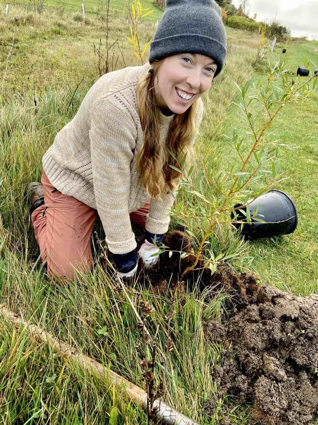 A woman planting a young tree in a grassy field, wearing a beanie, sweater, and pants, smiling at the camera.