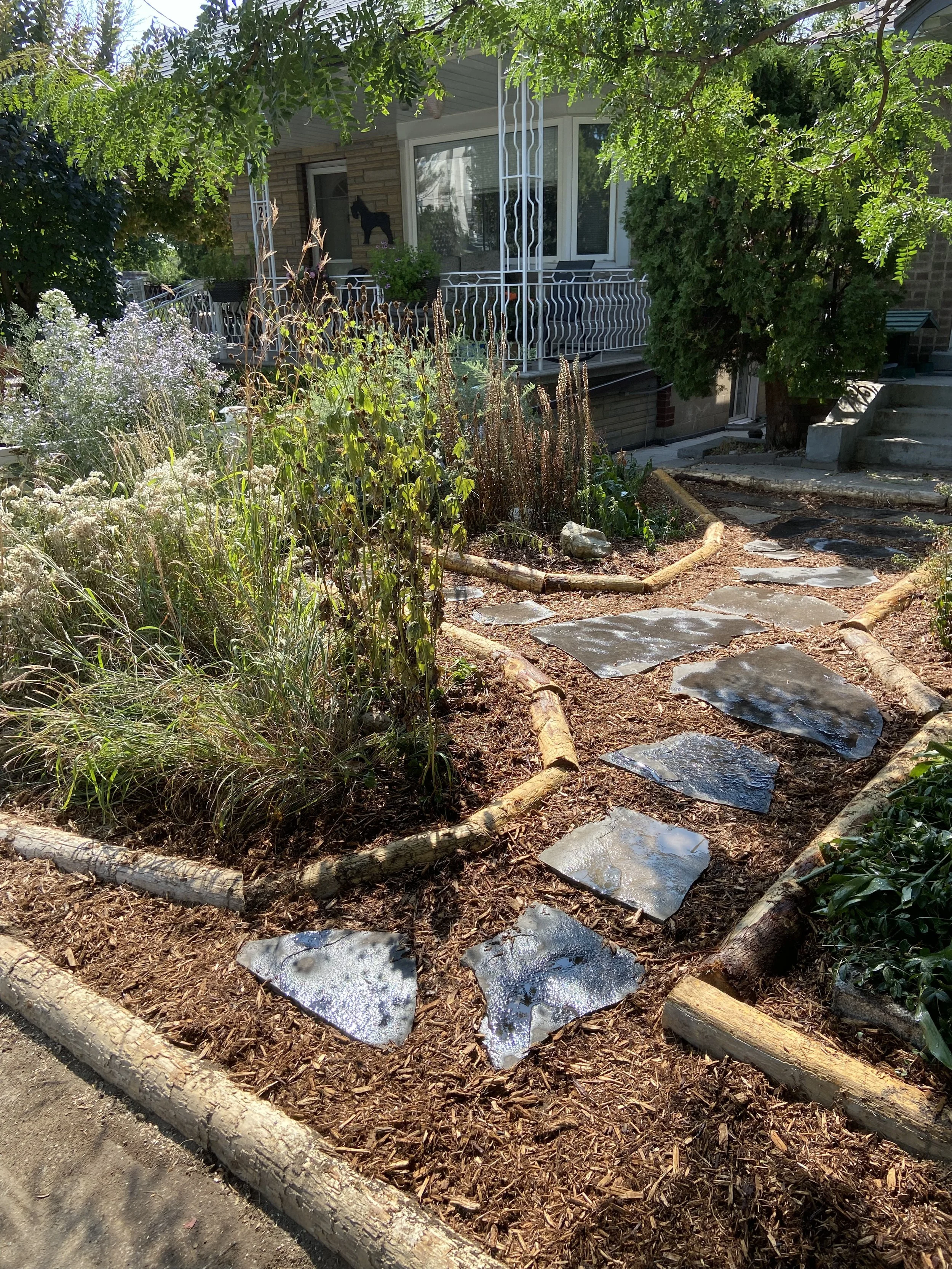 A garden pathway made of irregularly shaped stone slabs, bordered by wood logs, leading through a garden with plants, mulch, and trees to a house porch with railing and steps.