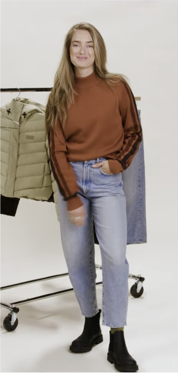 Young woman with long blonde hair smiling, wearing a brown sweatshirt, light-wash jeans, and black ankle boots, posing in a studio setting.