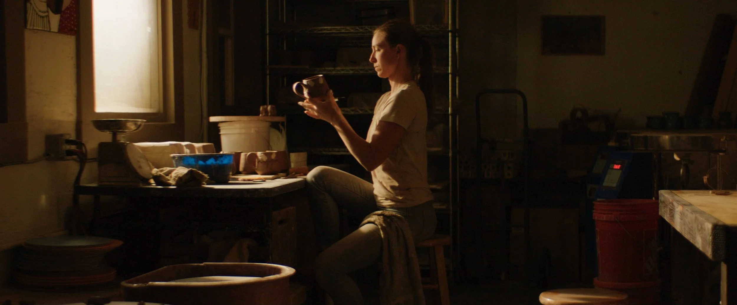 A woman sits on a stool in a dimly lit room, holding a mug and looking at it, with warm sunlight coming through a window behind her.