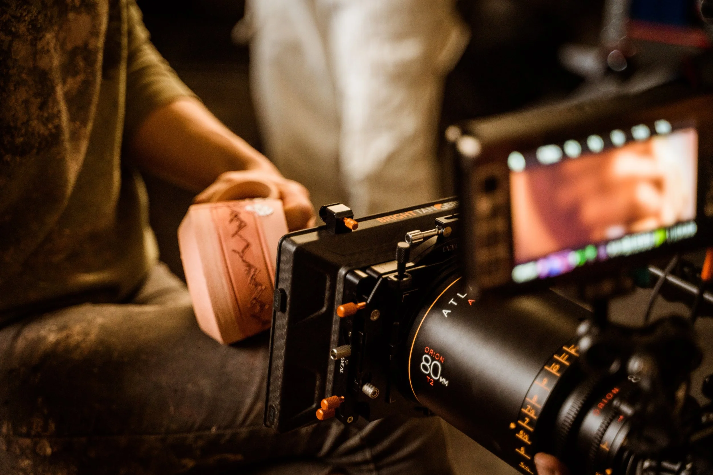 Close-up of a professional camera with a large lens, mounted on a tripod or stabilizer, with a monitor in the background displaying a scene being filmed.