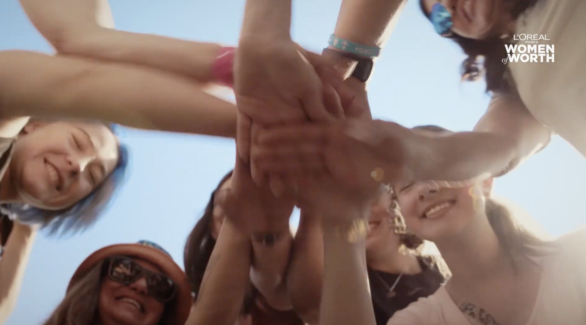 A group of smiling women stacking their hands together in a circle, taken from below with a clear blue sky in the background.