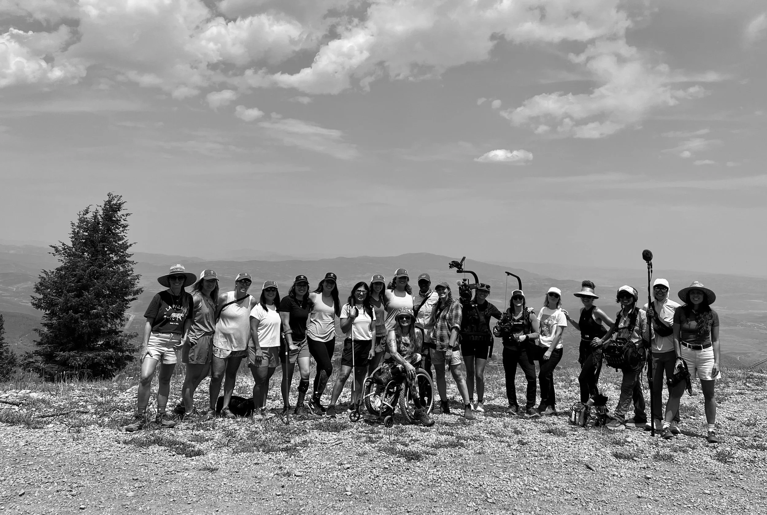 A group of people outdoors on a hillside with mountains in the background, some wearing hats and sunglasses. They include children and adults, some with wheelchairs and cameras, posing for a photo under a partly cloudy sky.