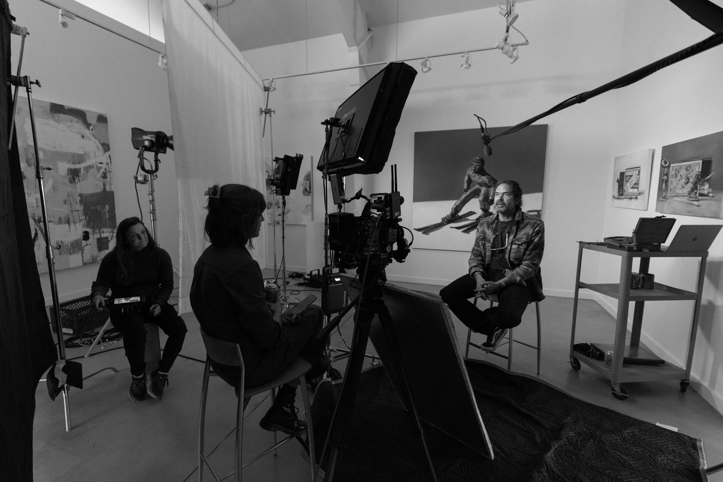 A behind-the-scenes look at a video or photo shoot in an art gallery. A man with curly hair and a beard is seated in front of a camera, speaking or being interviewed. Two women are present, one sitting on a chair and the other operating the camera. The gallery has artwork displayed on the walls, and professional lighting and audio equipment are set up around the scene.