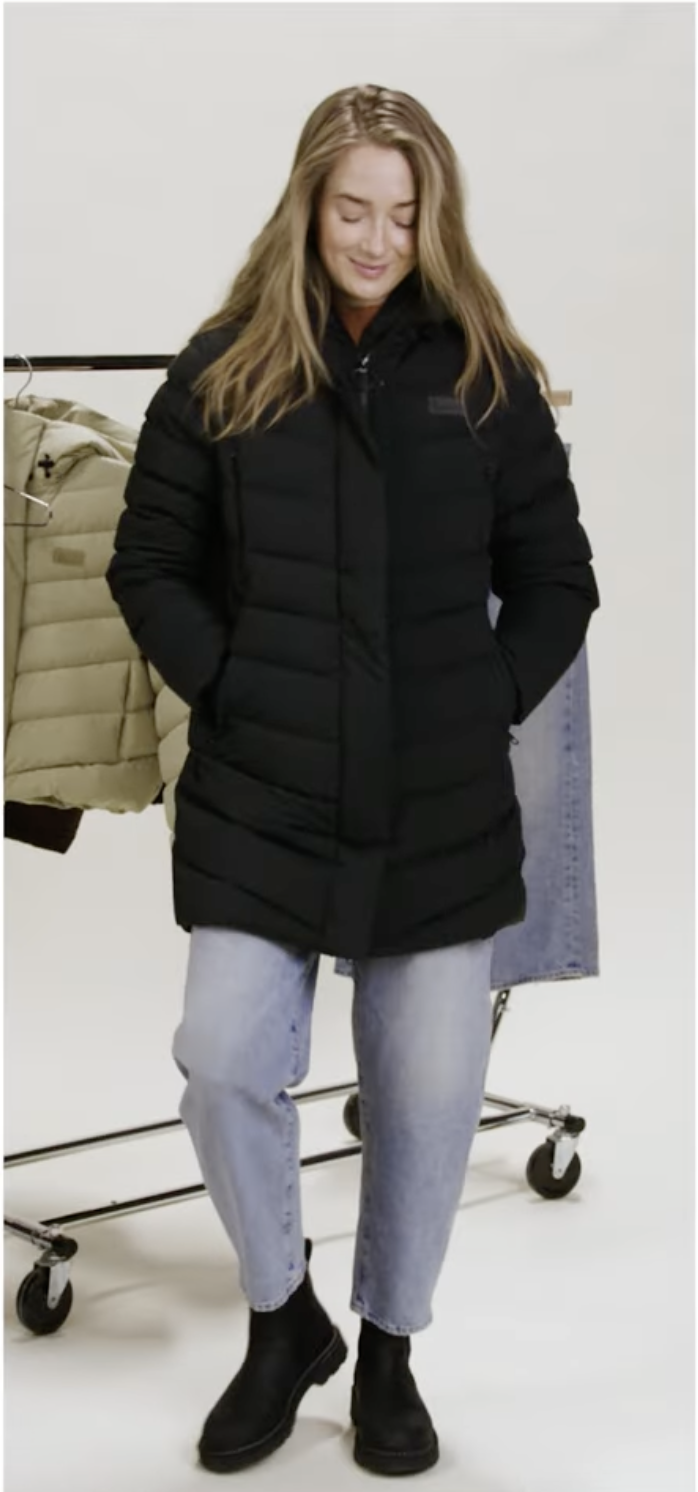 Woman wearing a black winter jacket, light blue jeans, and black ankle boots, standing with hands in pockets in a clothing store or studio.