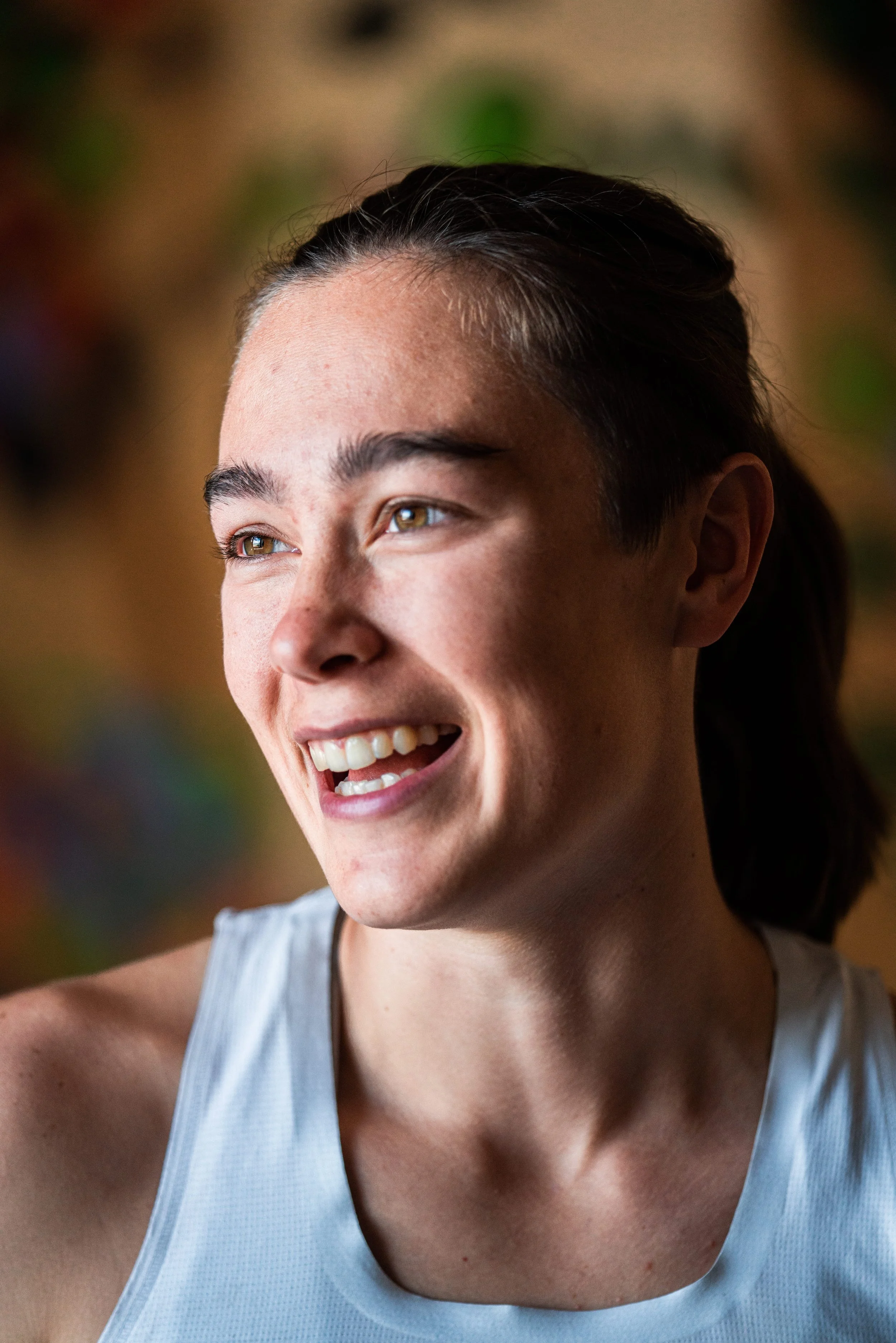 Close-up of a smiling woman with dark hair tied back, wearing a light blue athletic top, in a brightly lit environment.